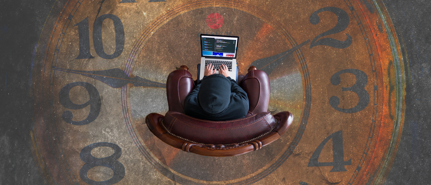 An overhead image of a man in a black hoodie sitting on a chair with a laptop, over a large painting of a clock on the floor.