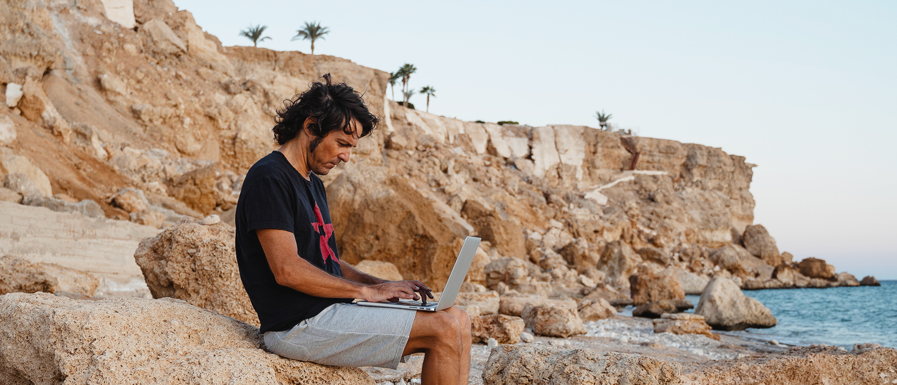 Man sitting on rock at the beach while using a laptop
