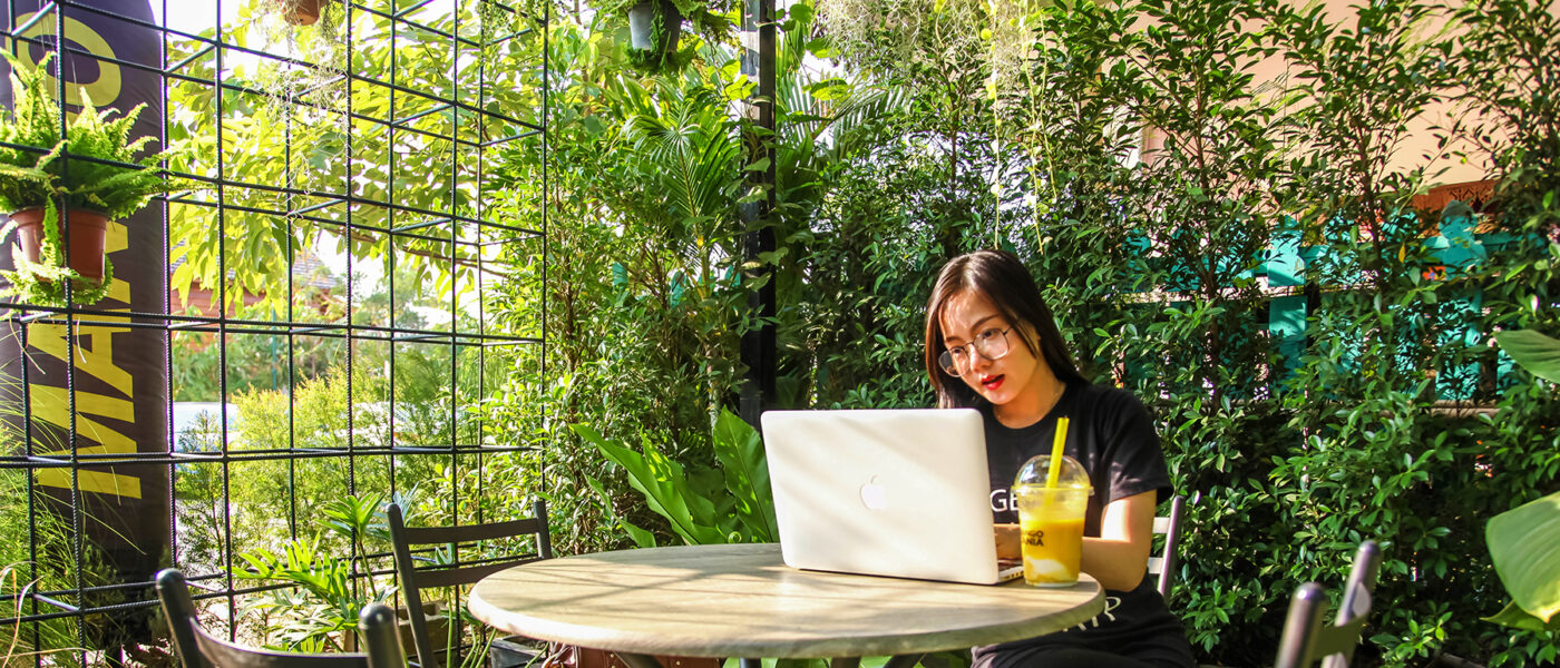 Woman sitting on chair while using laptop in a glass room surrounded by green plants. It's sunny outside.
